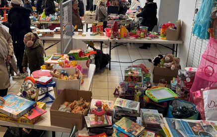 Un marché aux puces intérieur animé proposant des tables remplies de divers jouets, jeux et articles pour enfants à vendre, avec des acheteurs parcourant les marchandises colorées.
